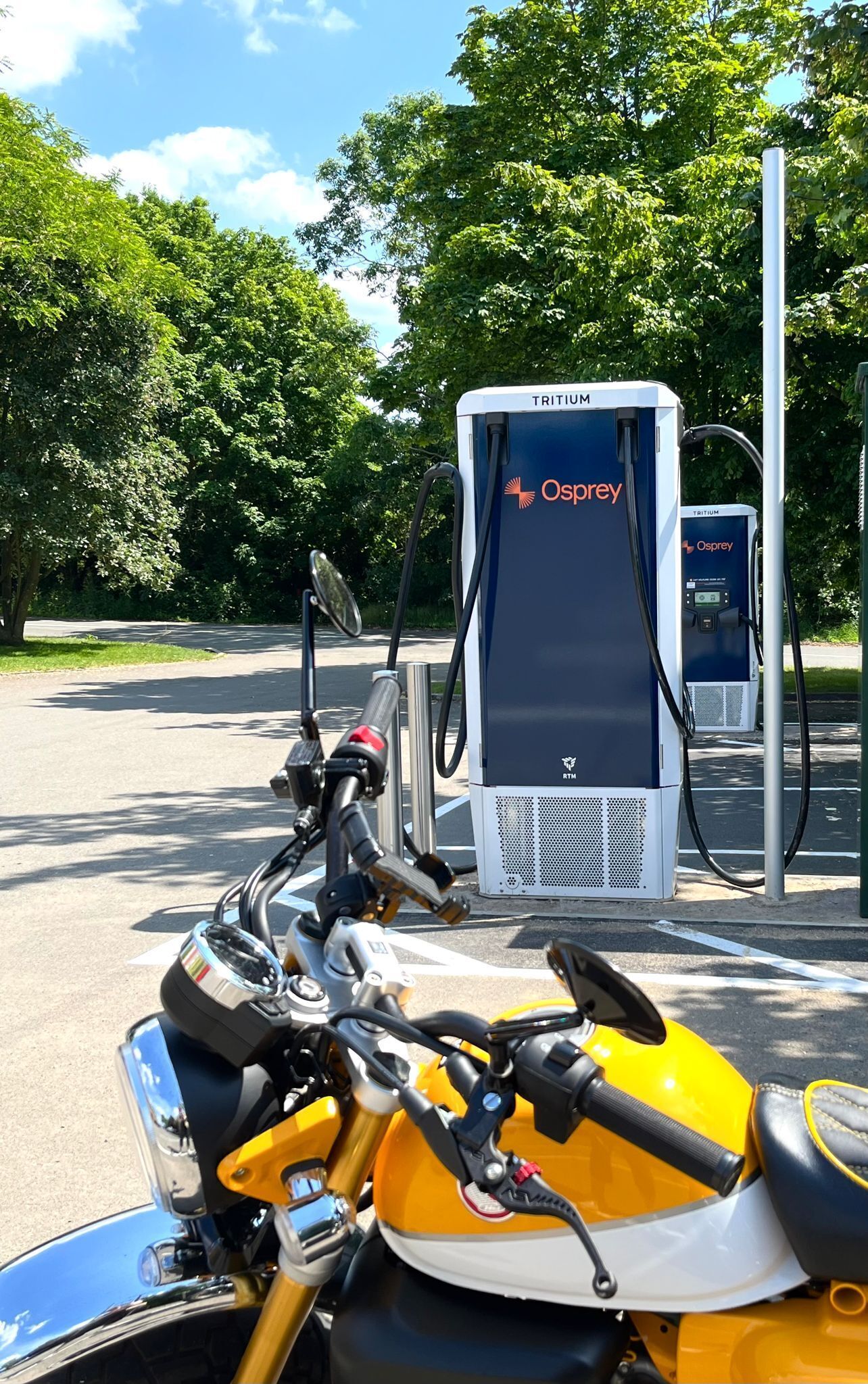 Een gele motorfiets geparkeerd bij een Tritium Osprey laadstation voor elektrische voertuigen.