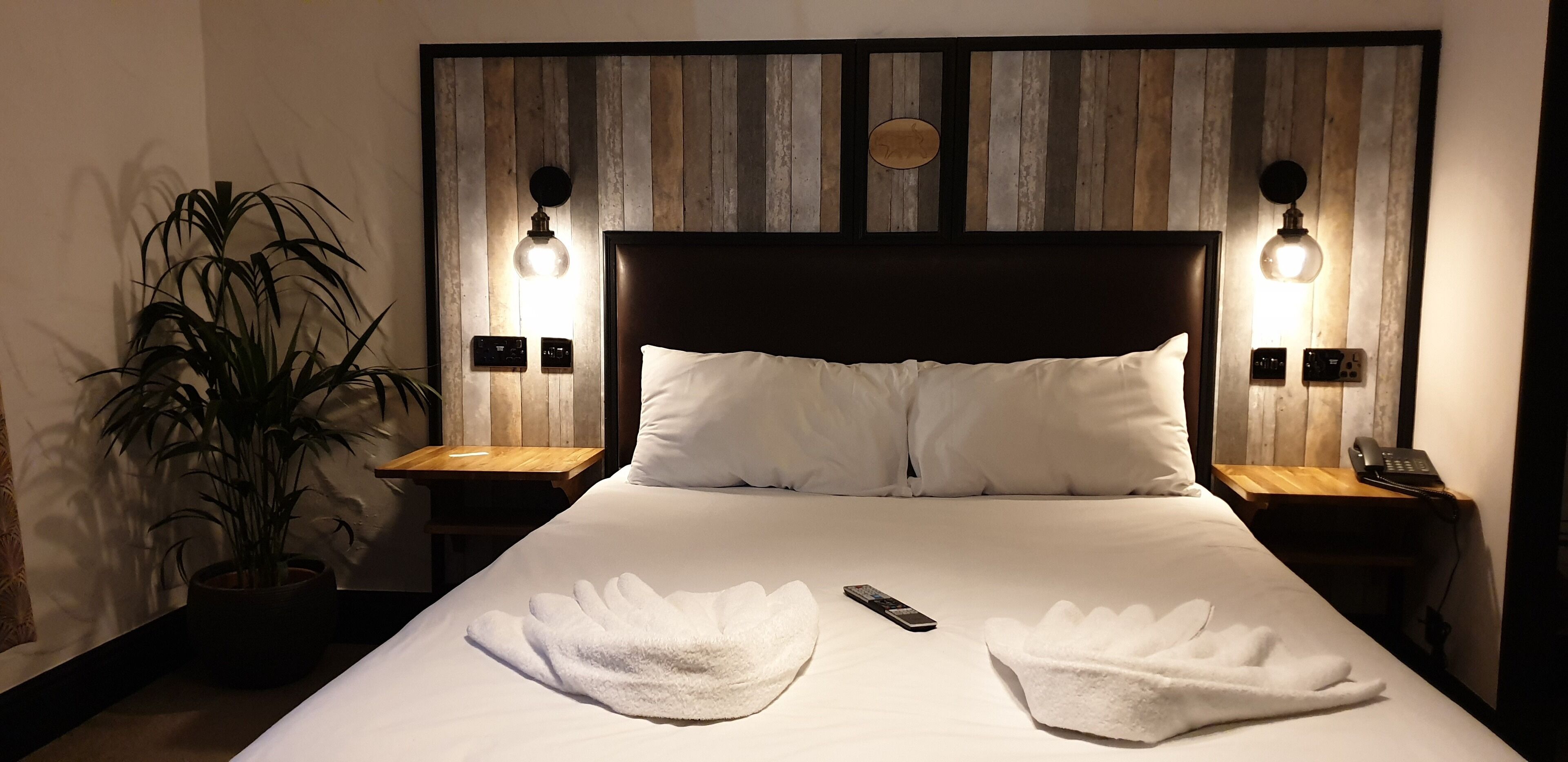 A neatly made bed with white linens, two bedside lamps, a potted plant, and a bedside phone on a wooden nightstand.