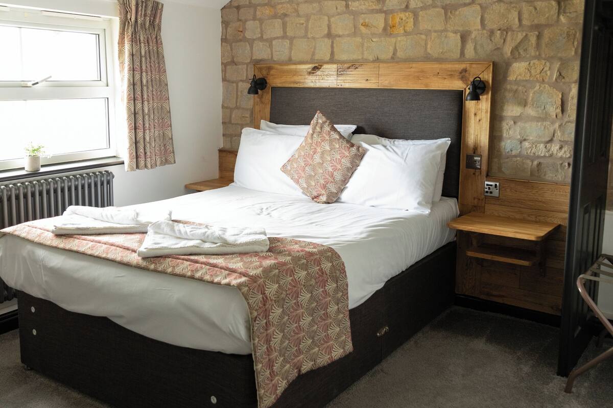 A bedroom with a stone wall, a wooden headboard, a bed with white linens, a bedside table, and a window with curtains.
