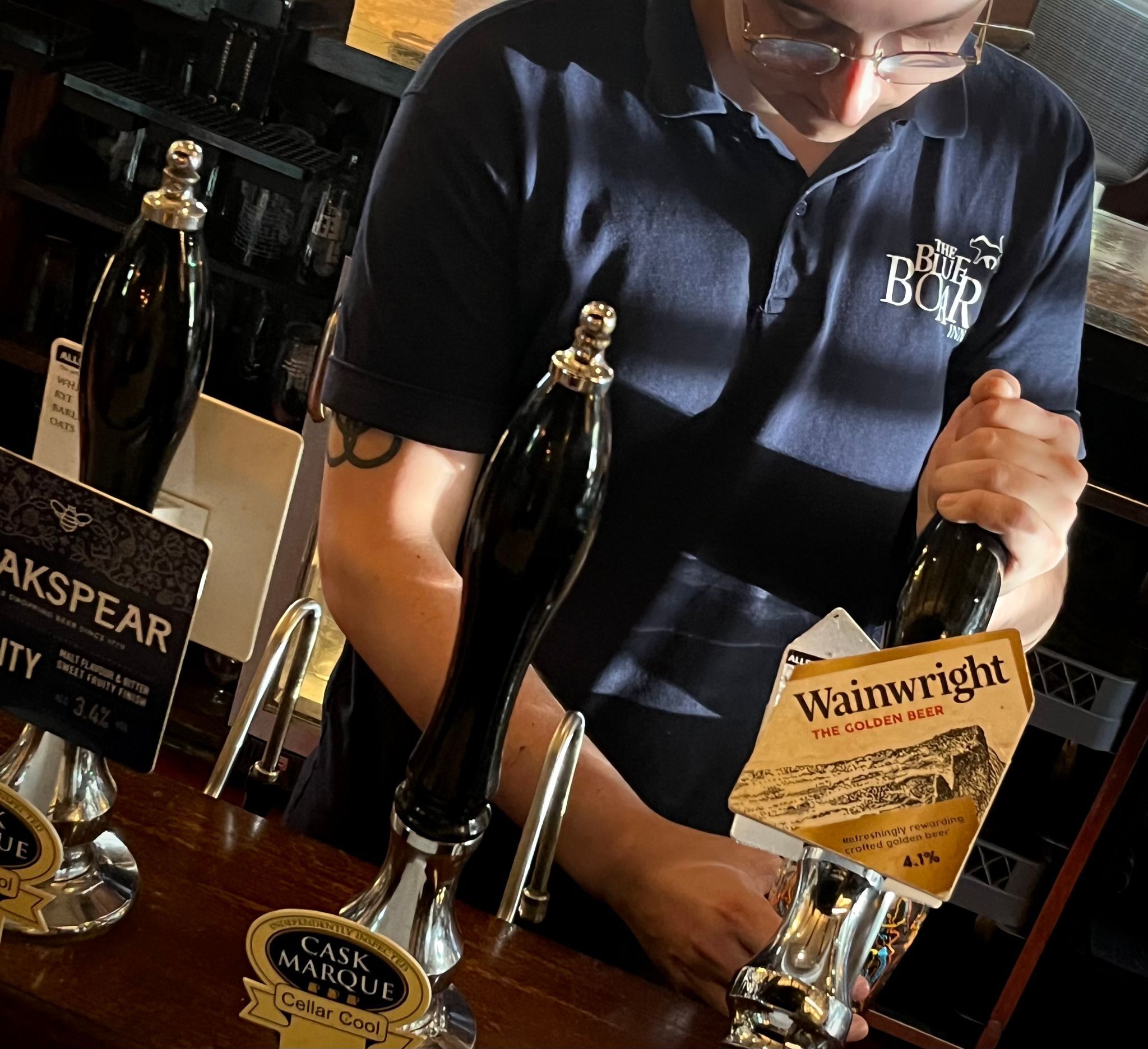 Un cantinero detrás de una barra con grifos etiquetados 'Cask Marque' y 'Wainwright'.