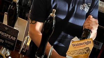 Un cantinero detrás de una barra con grifos etiquetados 'Cask Marque' y 'Wainwright'.
