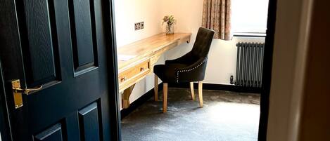 A room with a dark wooden door, a desk with a lamp, a chair, and a radiator.