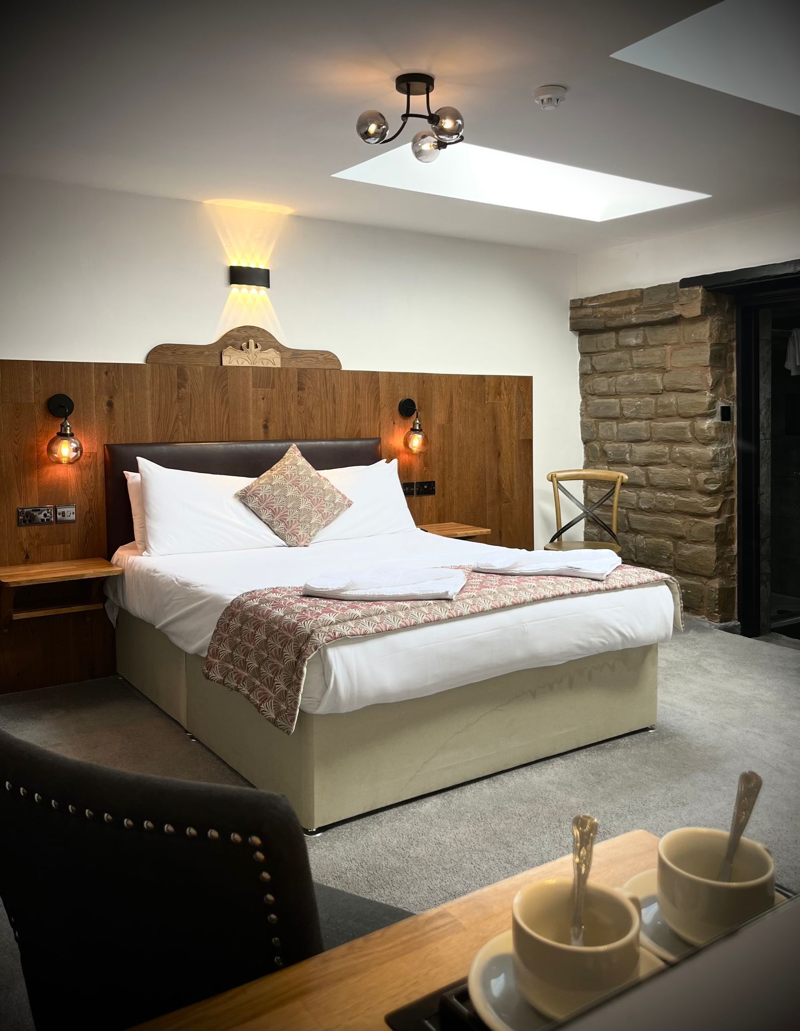 A neatly made bed with a wooden headboard, two bedside lamps, a stone wall, and a skylight above.