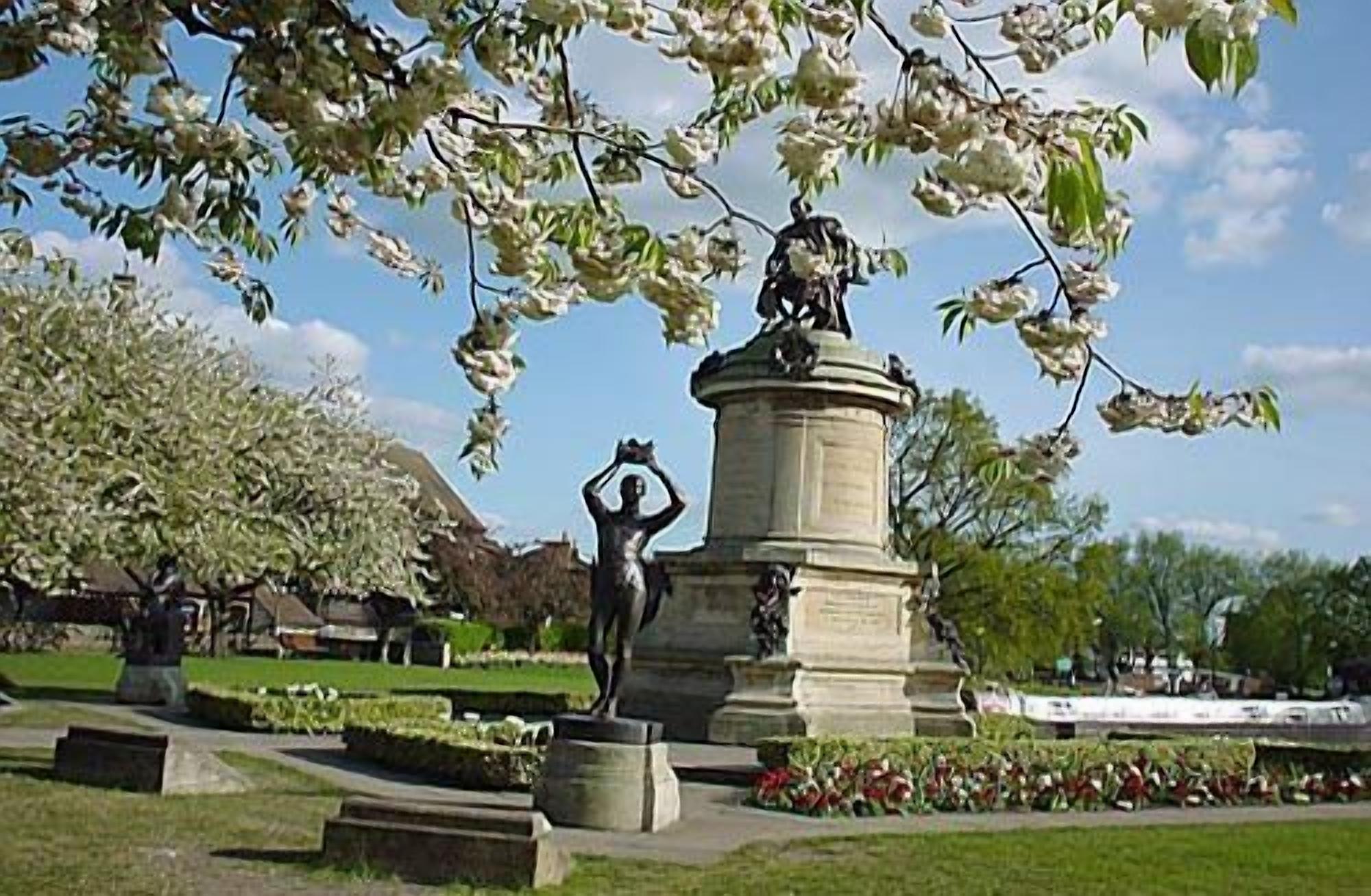 A park with a statue of a man holding a sword, surrounded by well-maintained grass and flowering trees.