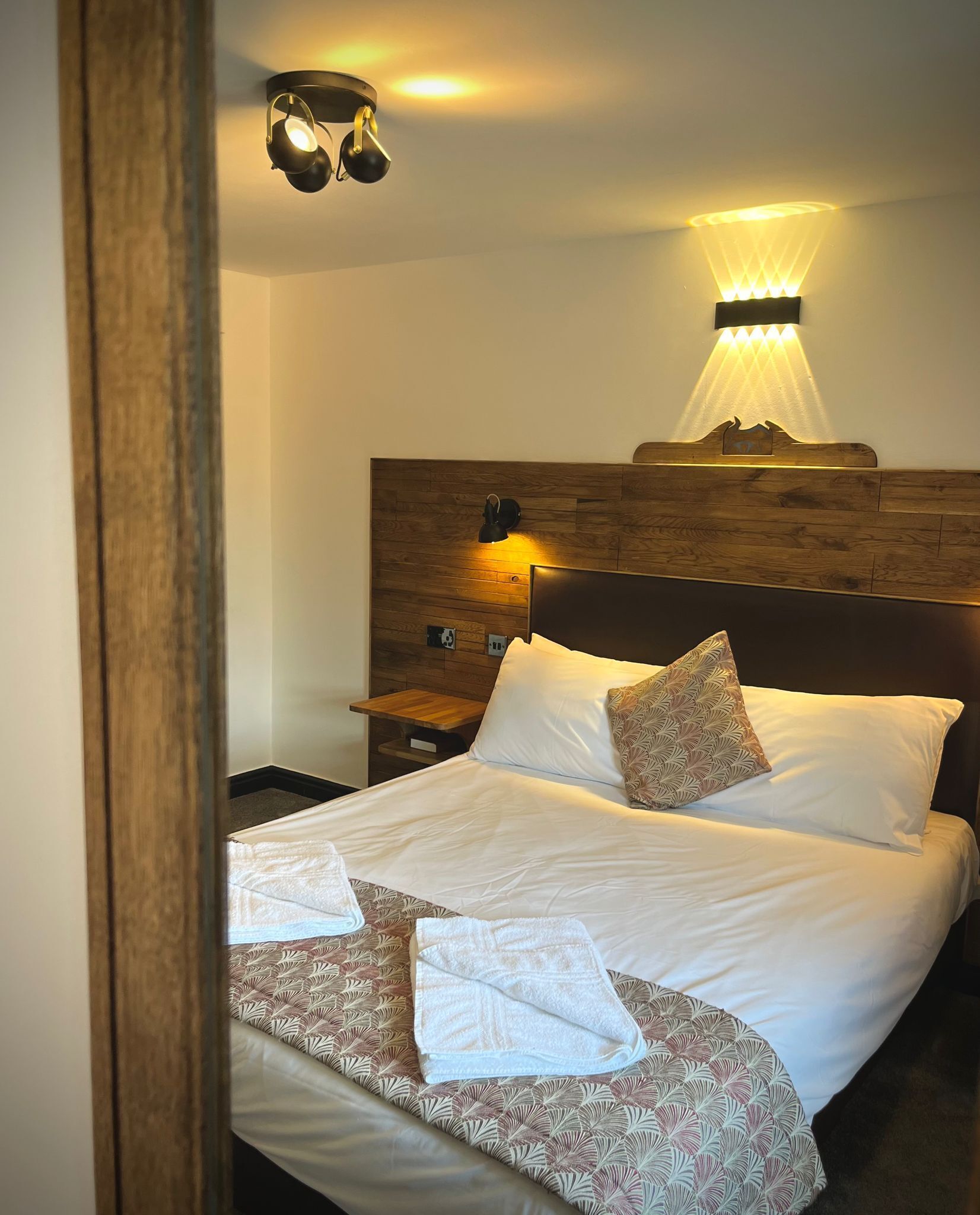 A neatly made bed with white linens and patterned pillows, a wooden headboard, and wall-mounted lighting.