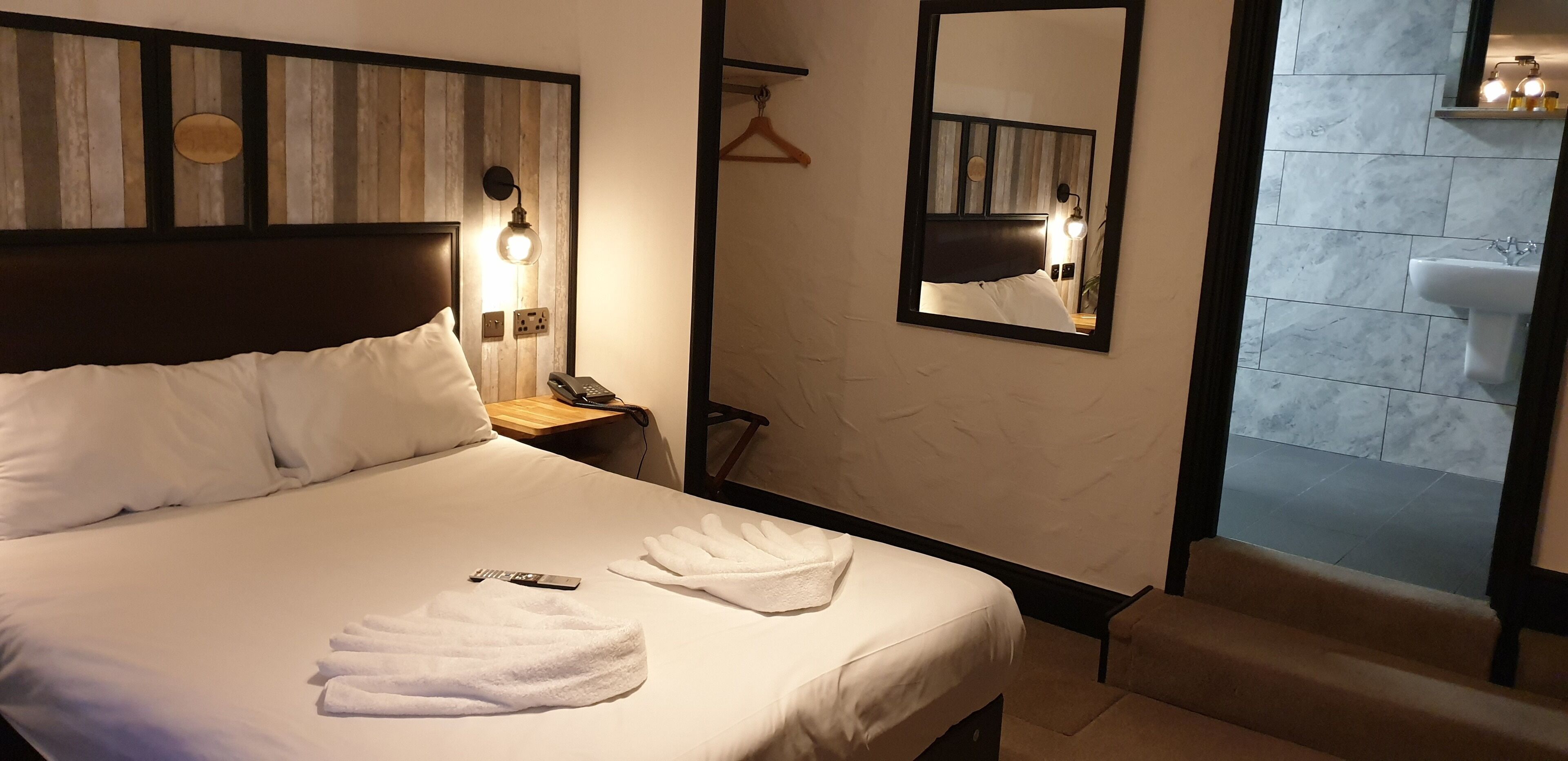 Ein Hotelzimmer mit einem großen Bett, einem Nachttisch mit Lampe, einem Spiegel und einem durch eine offene Tür einsehbaren Badezimmer.