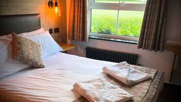 A neatly made bed with white linens, a patterned pillow, and a folded towel on a bed with a patterned bedspread. A wall-mounted light fixture and a window with curtains are visible.