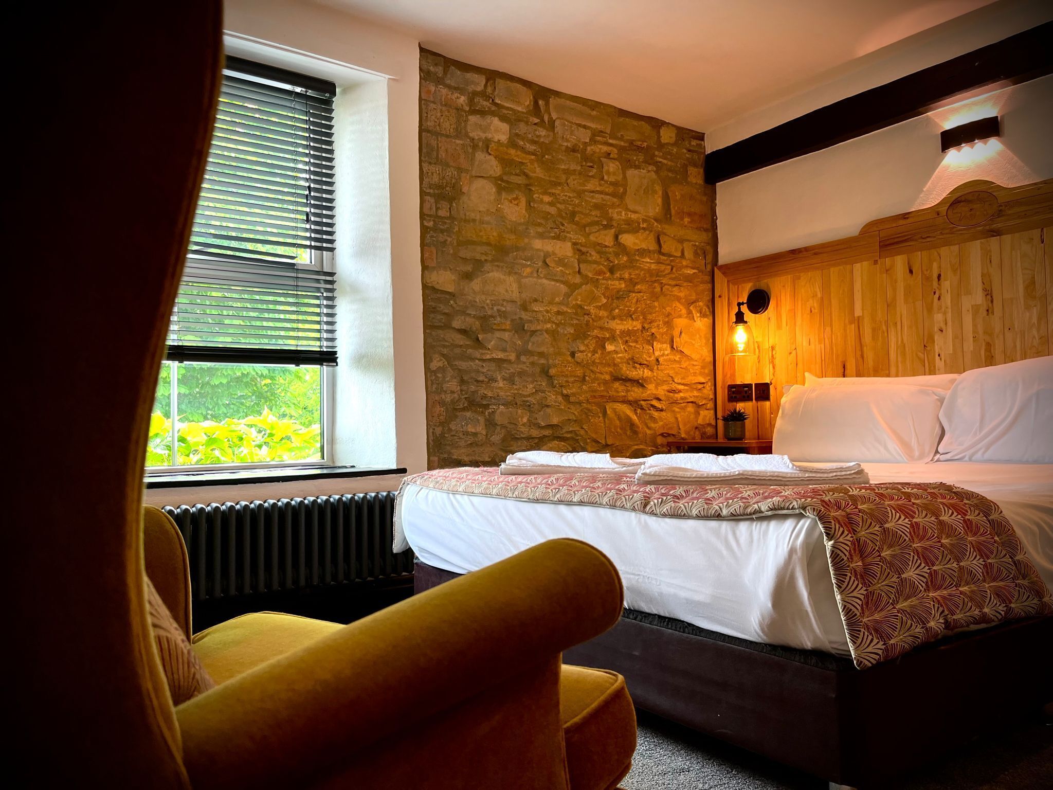 Een slaapkamer met een stenen muur, een bed met witte lakens, een houten hoofdbord en een raam met jaloezieën.