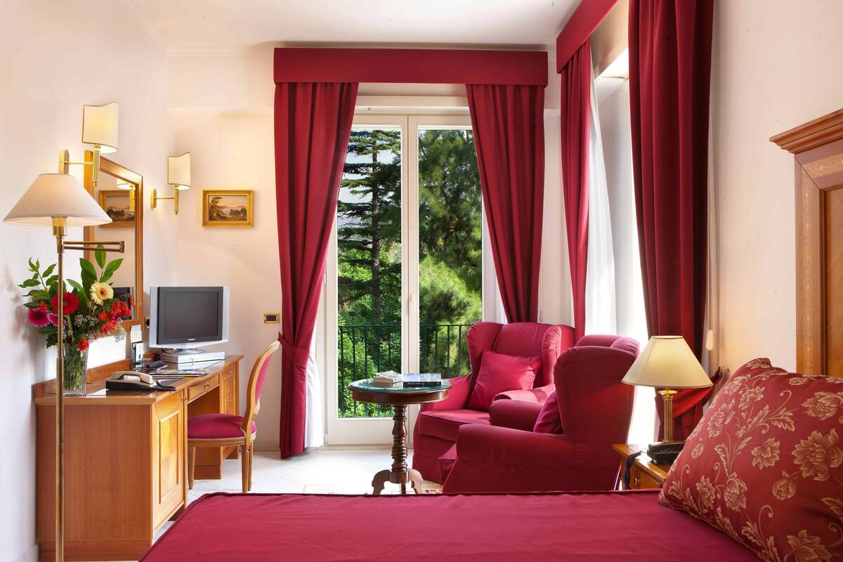 Una camera d'albergo con un letto, una scrivania con un computer, una sedia, una poltrona rossa, un tavolino e una finestra con tende.