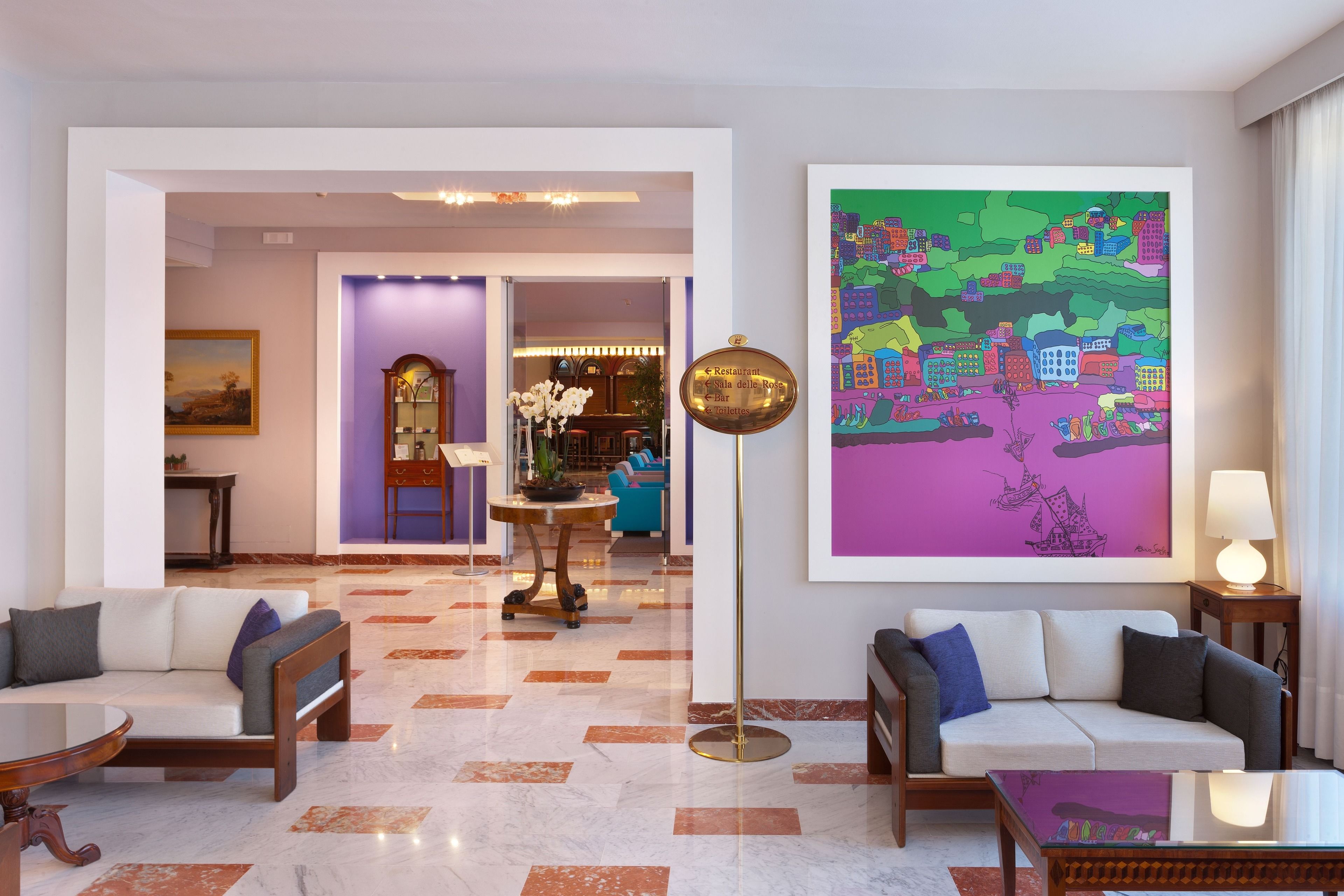 Un hall d’hôtel avec deux canapés, une lampe sur pied, une grande fresque colorée et un comptoir de réception.