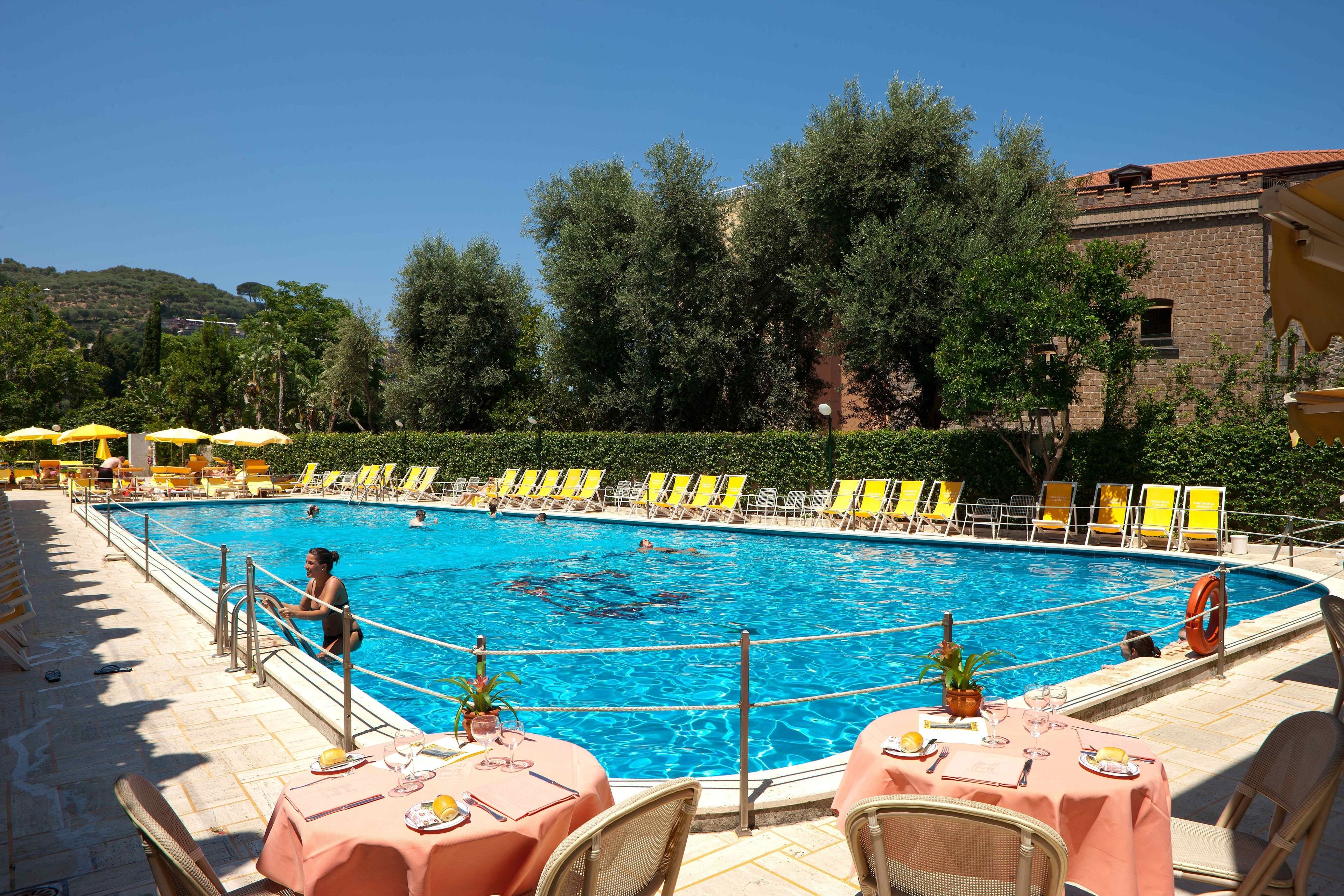 A swimming pool with yellow chairs and tables, surrounded by greenery and a building.