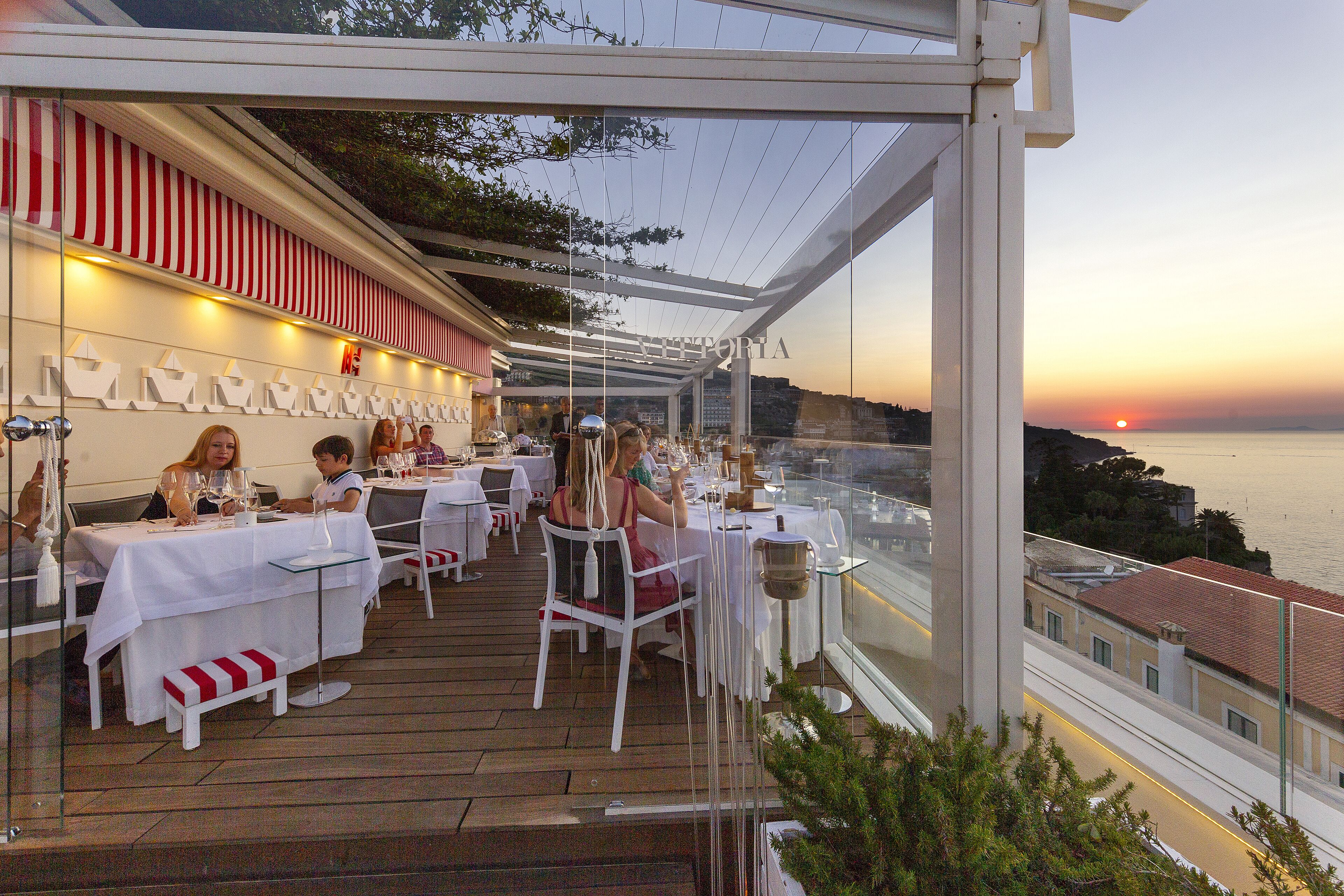 An outdoor dining area with tables and chairs, overlooking a scenic view of the sunset and the sea.
