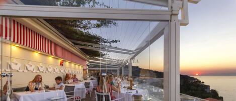 An outdoor dining area with tables and chairs, overlooking a scenic view of the sunset and the sea.