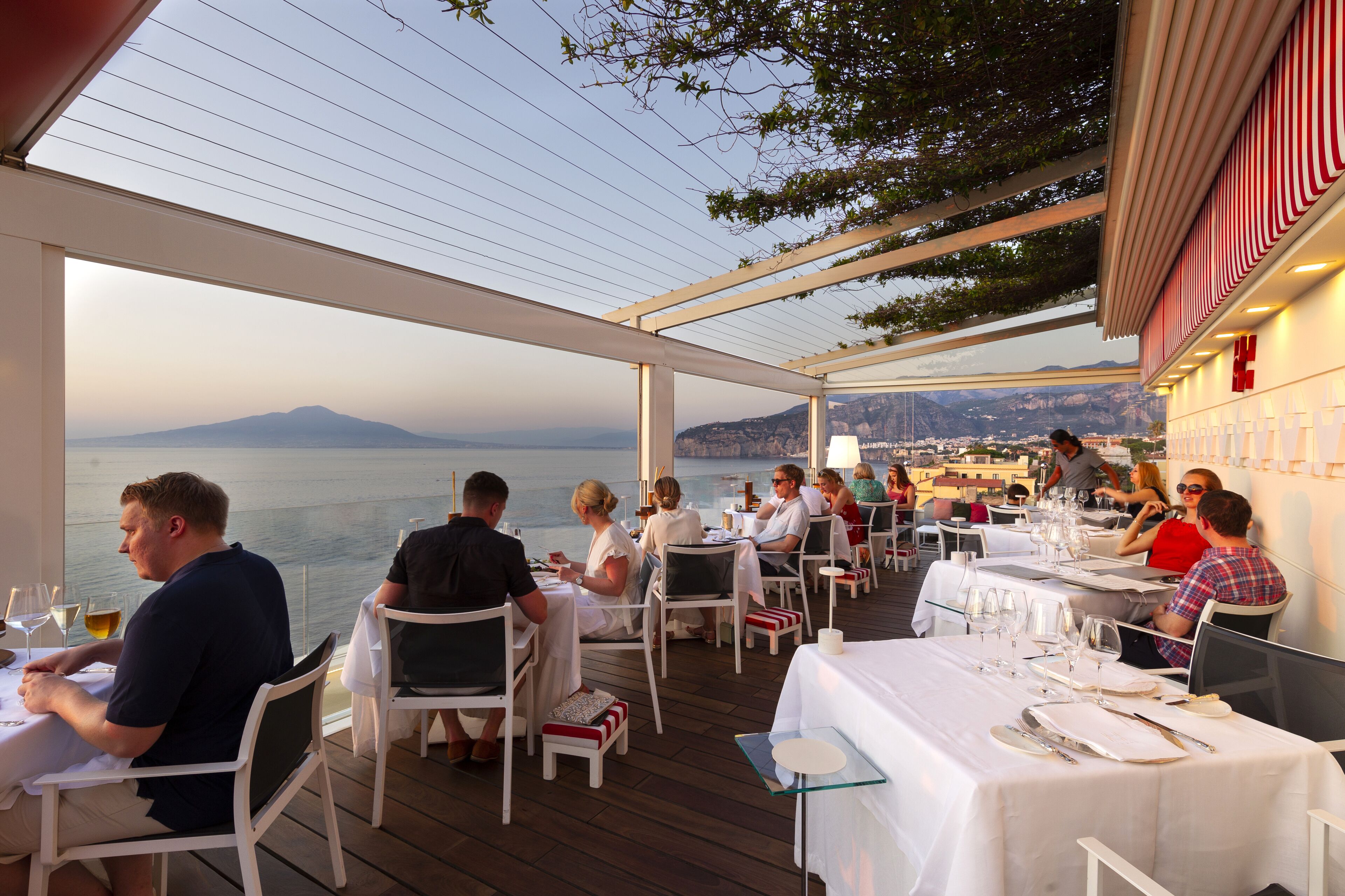 An outdoor dining area with tables and chairs, overlooking a body of water and distant mountains.