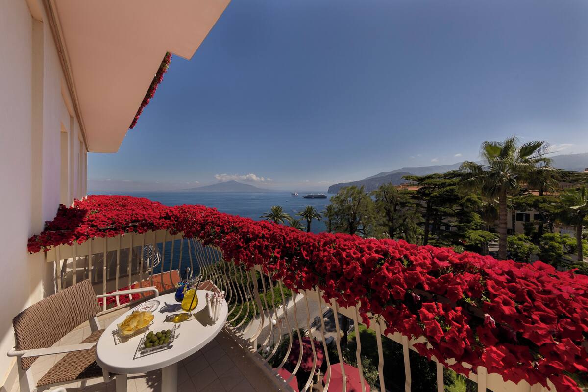 Un balcone con fiori rossi, un tavolo imbandito e vista sul mare e sulle montagne.