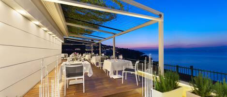 Un área de comedor al aire libre con mesas y sillas, una terraza de madera y vista al mar.