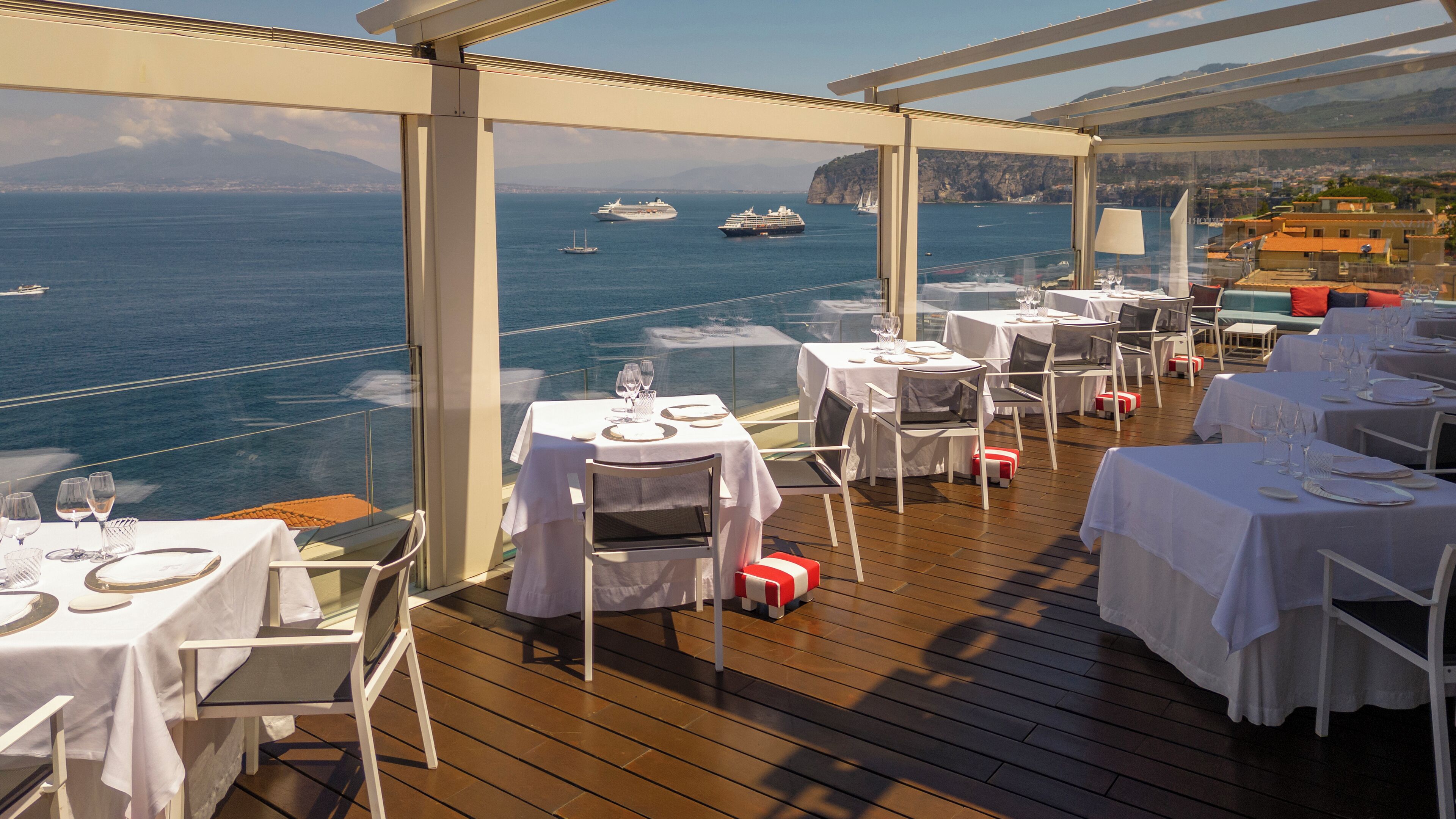 Comedor con mesas dispuestas para una comida, con vista a un cuerpo de agua y barcos.