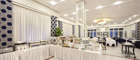 Un salón de banquetes con una larga mesa dispuesta para un bufé, rodeado de sillas y mesas.