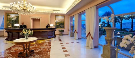 A hotel lobby with a reception desk, a chandelier, a large window with a view of palm trees, and a marble floor.