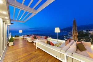 A deck with white sofas and tables, a lamp, and a view of the sea.