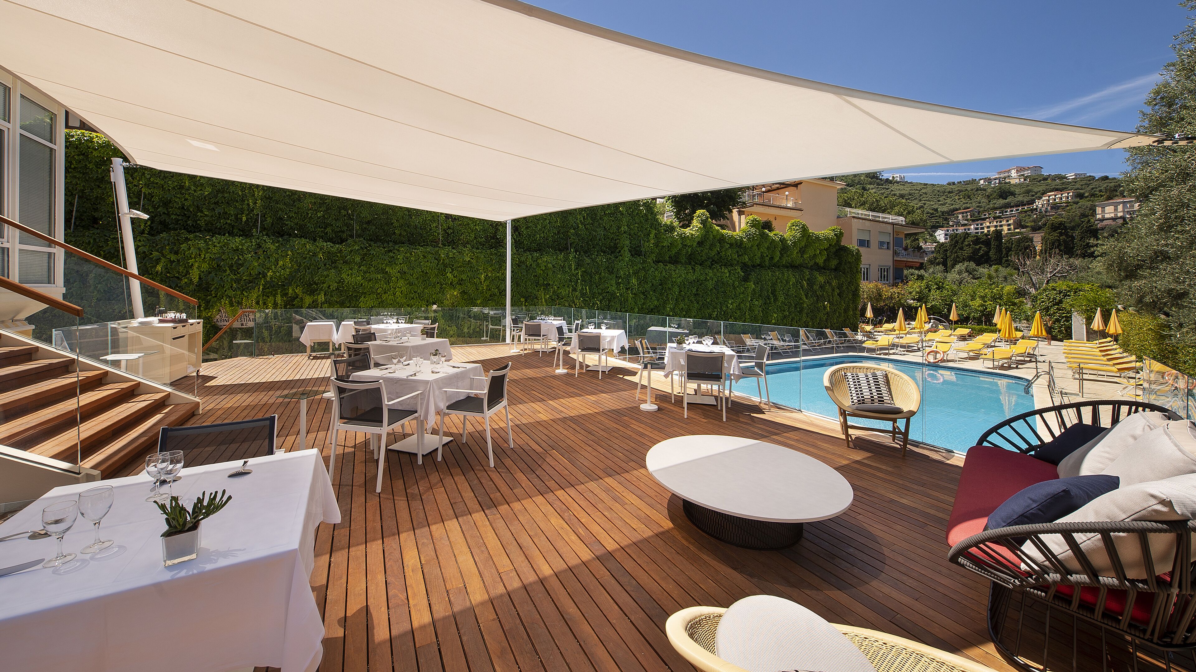 A rooftop terrace with wooden flooring, white tables and chairs, a pool, and a view of a hillside.