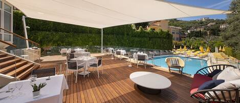 Terraza en la azotea con suelo de madera, mesas y sillas blancas, una piscina y vistas a una colina.