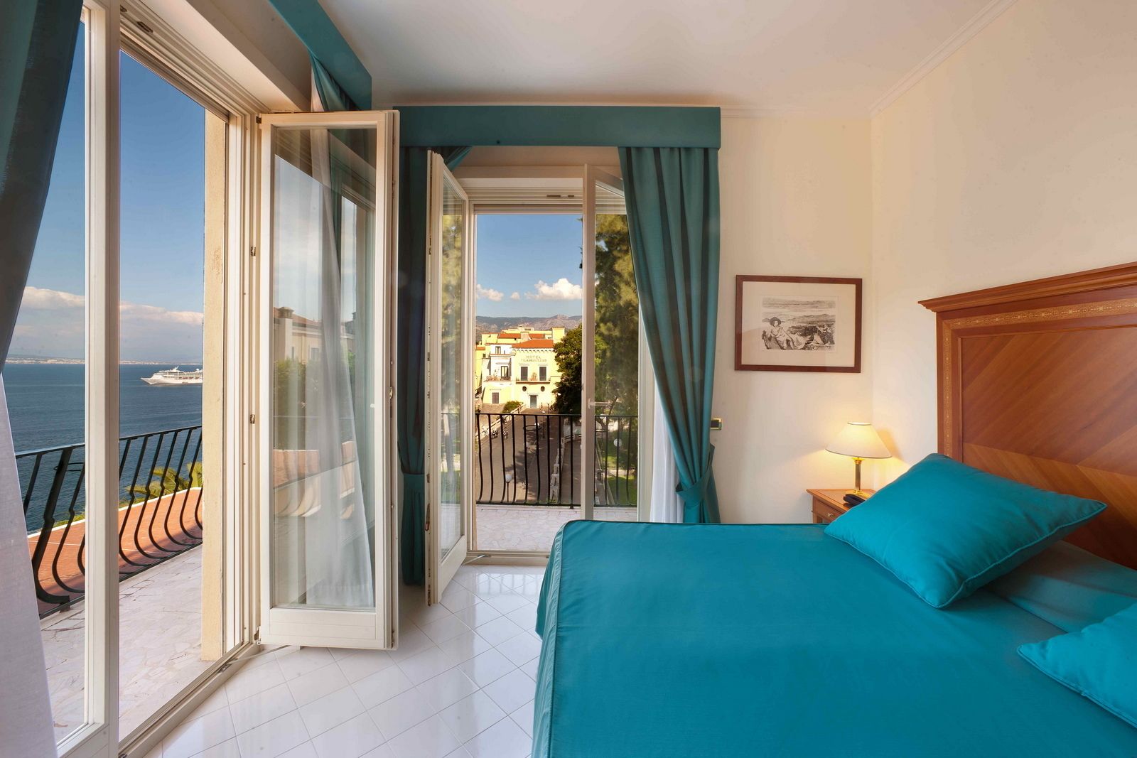 A room with a bed, a nightstand, a lamp, and a view of the sea and buildings through an open door.