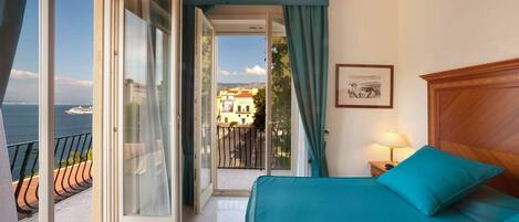 Una habitación con cama, mesita de noche, lámpara y vistas al mar y edificios a través de una puerta abierta.