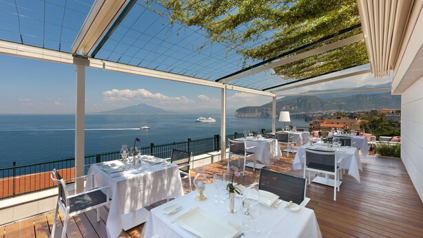 Un área de comedor al aire libre con mesas dispuestas para una comida, con vista a la costa y un barco a lo lejos.