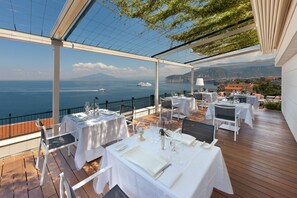 An outdoor dining area with tables set for a meal, overlooking a coastal view with a boat in the distance. - Continental (Sorrento)
