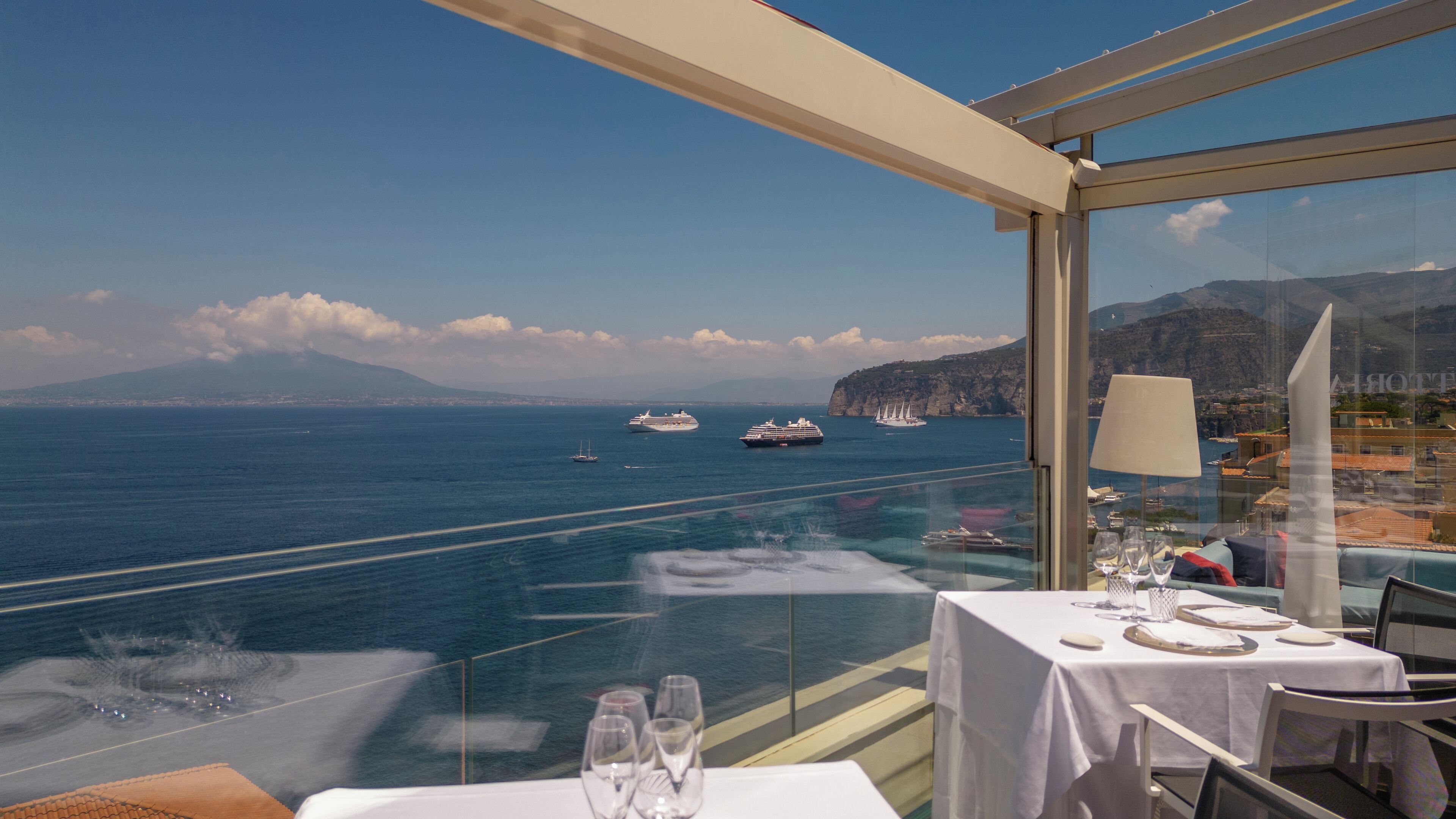 Comedor con una mesa dispuesta para dos, con vistas al mar, barcos y acantilados.