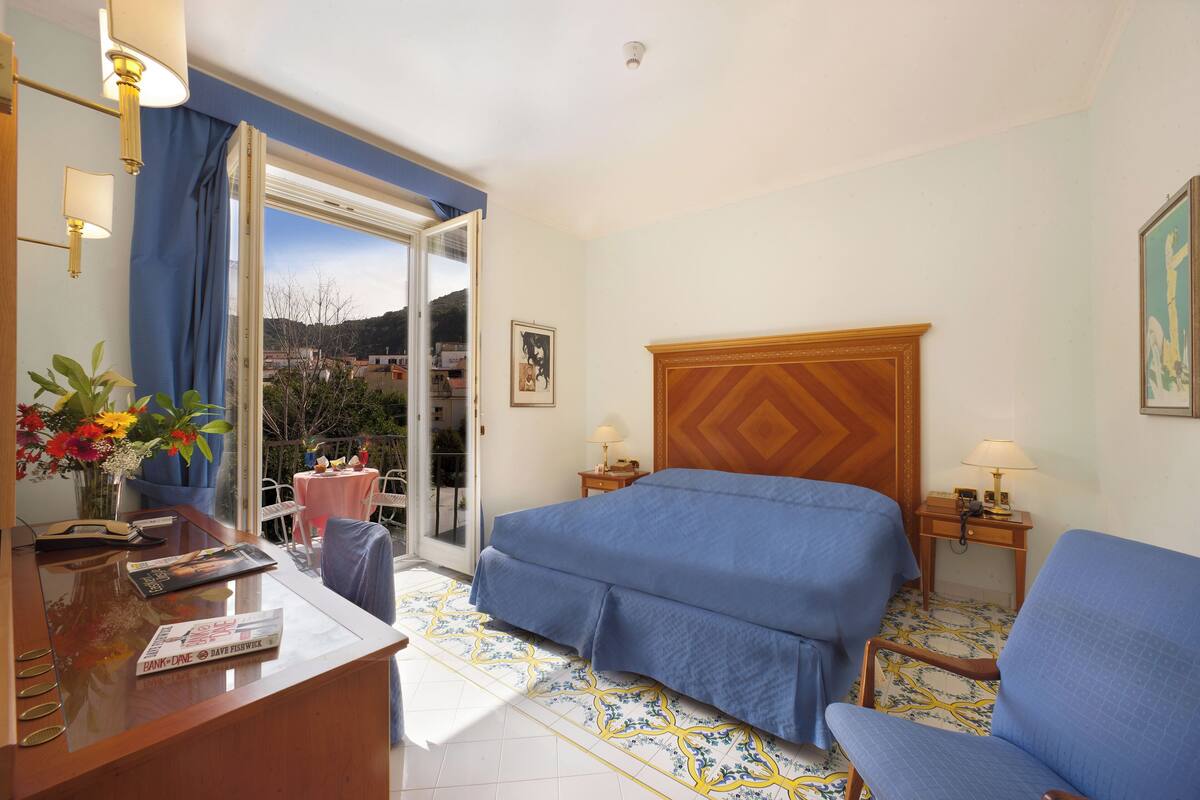 Una camera d'albergo con un letto, una scrivania con un computer, una sedia, un tavolino con una lampada e vista sull'esterno.