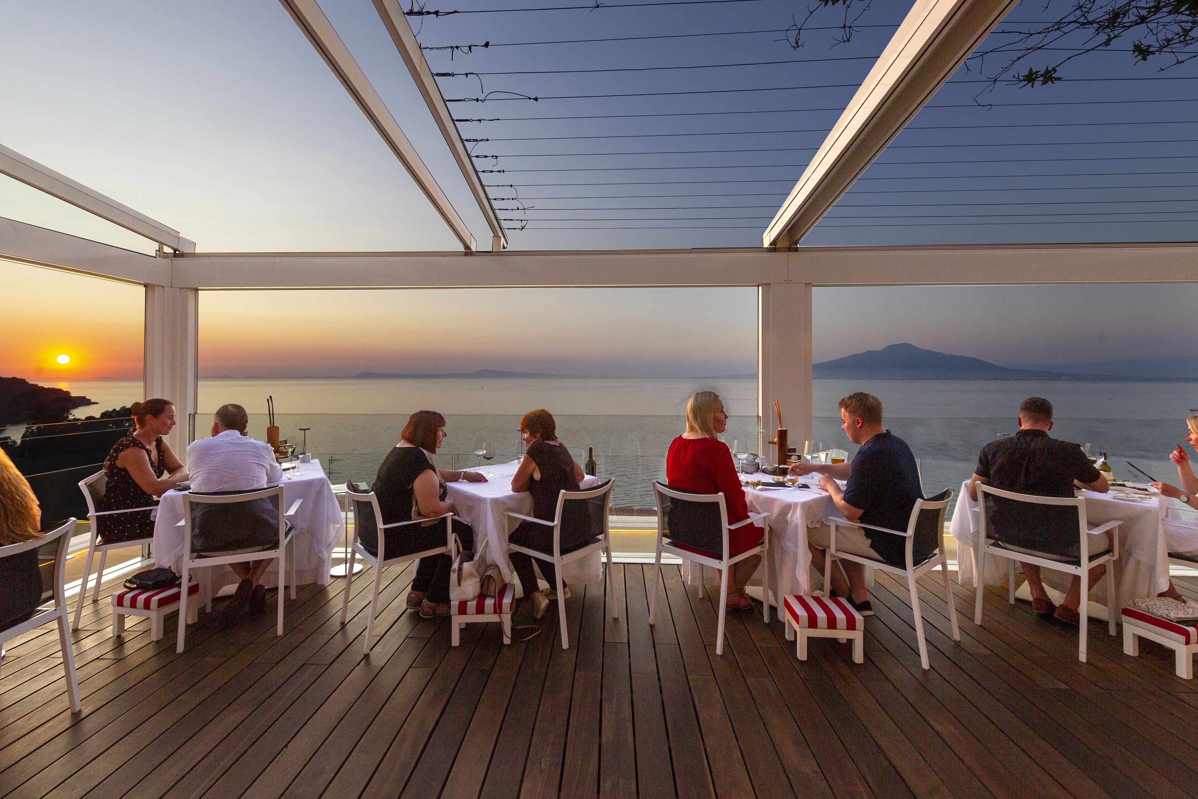 A dining area with tables and chairs, overlooking a body of water and a distant mountain range.
