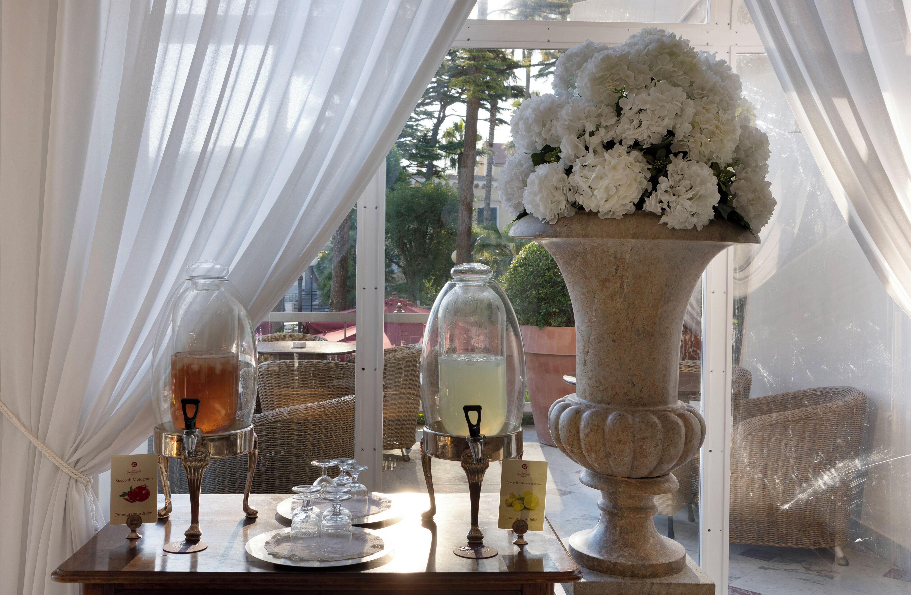 Una mesa con un botellero de vidrio, una jarra de vidrio, un florero con flores blancas y una silla de mimbre.