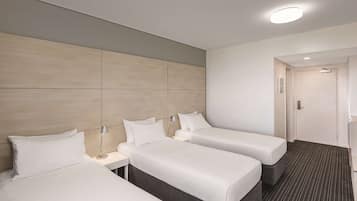 A hotel room with three single beds, a bedside table with a lamp, a modern light fixture, and a door leading to another room.