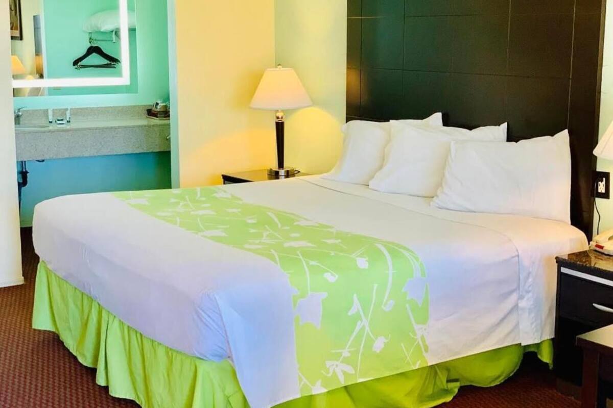 A hotel room with a large bed, a nightstand with a lamp, a mirror, and a small shelf with a hanger.