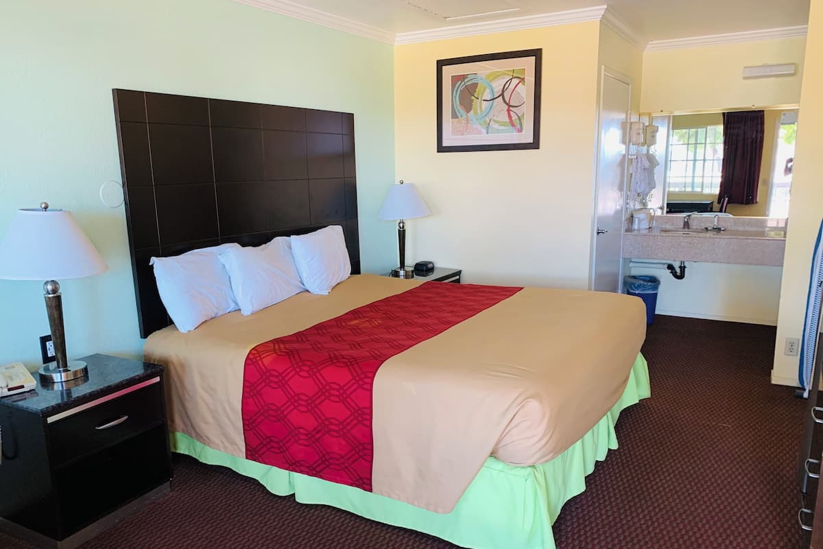 A hotel room with a large bed, two bedside tables, a nightstand, a bathroom with a sink and mirror, and a wall-mounted painting.