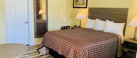 Habitación de hotel con cama, escritorio, silla, espejo, mesita de noche y lámpara.