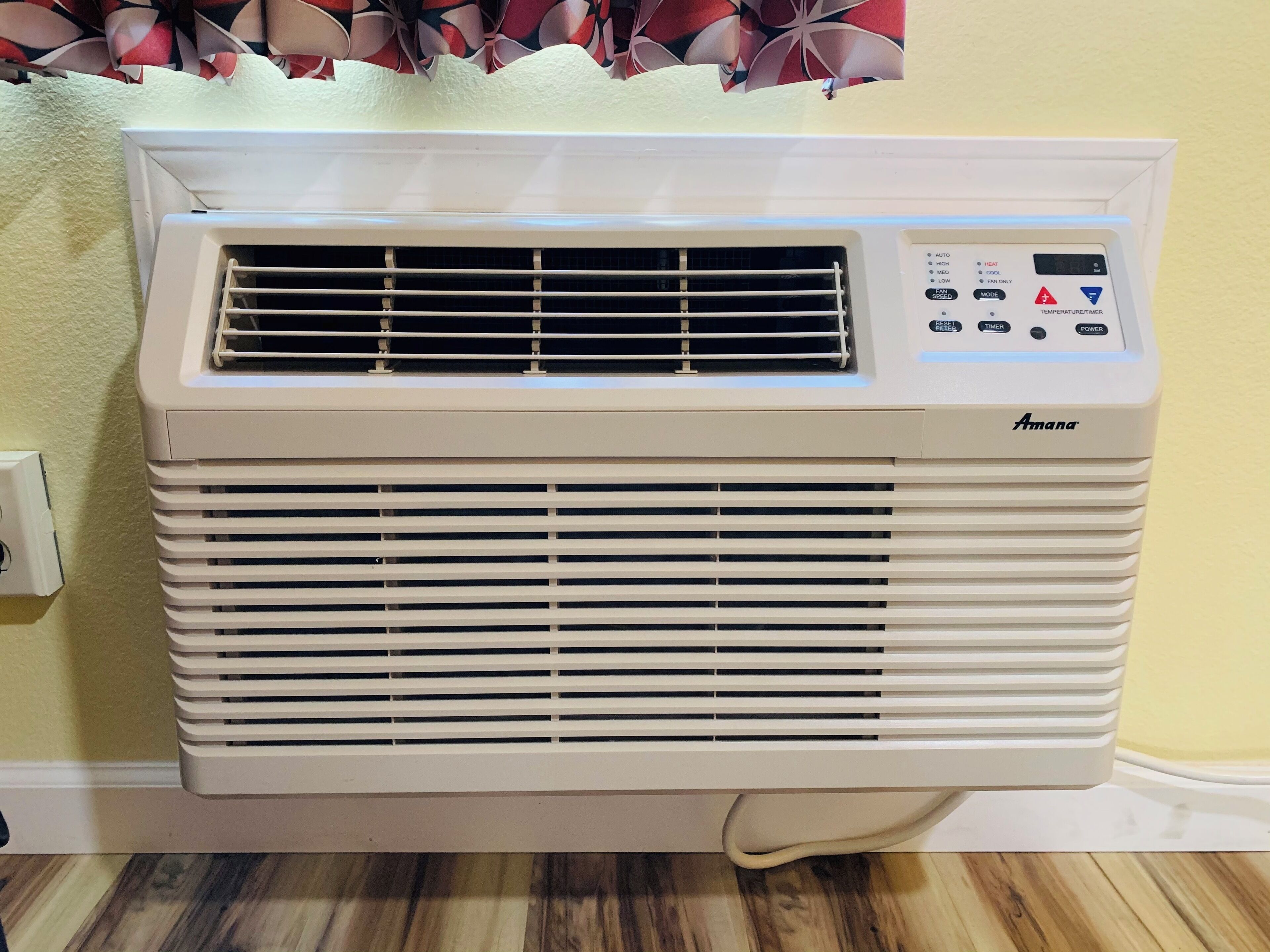 A white portable air conditioner with a control panel, placed against a wall with a patterned curtain.