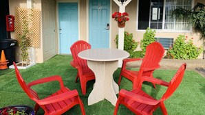 Una casa con puerta azul, un pequeño jardín de césped artificial y una mesa con cuatro sillas rojas.