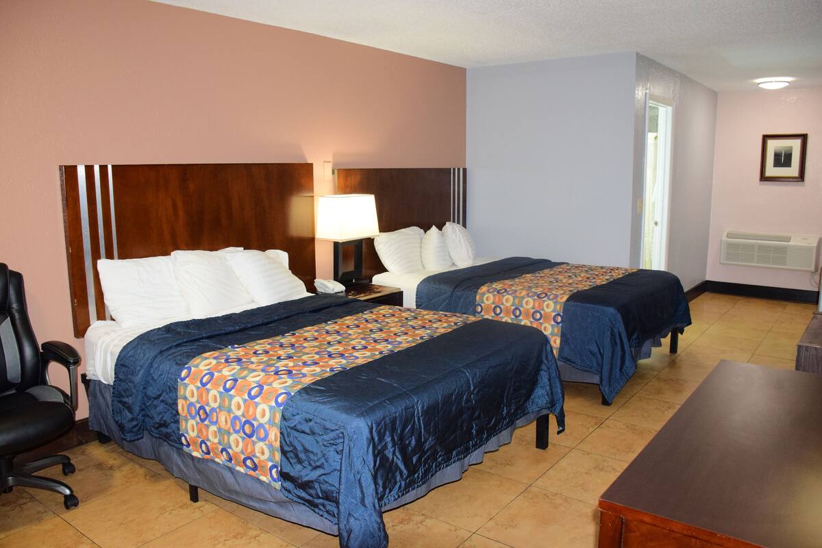 A hotel room with two beds, a desk, a chair, and a nightstand with a lamp.