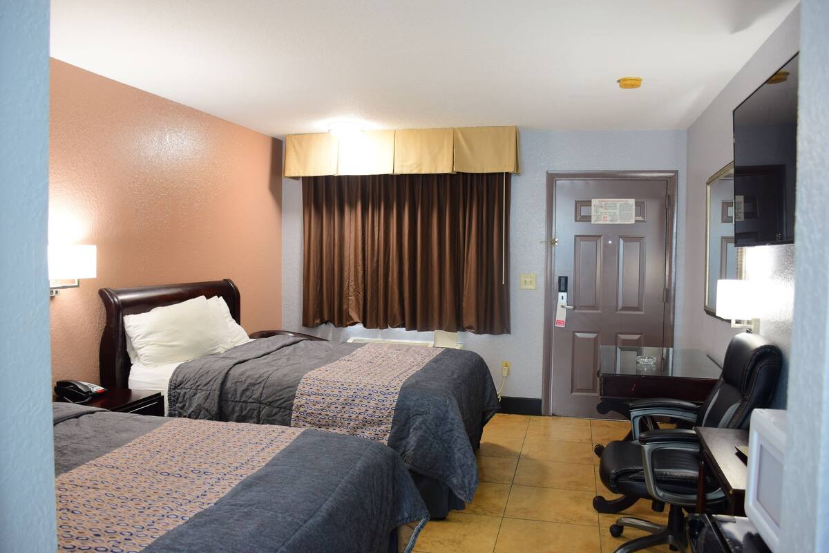 A hotel room with two beds, a desk, a chair, a TV, and a door with a sign.
