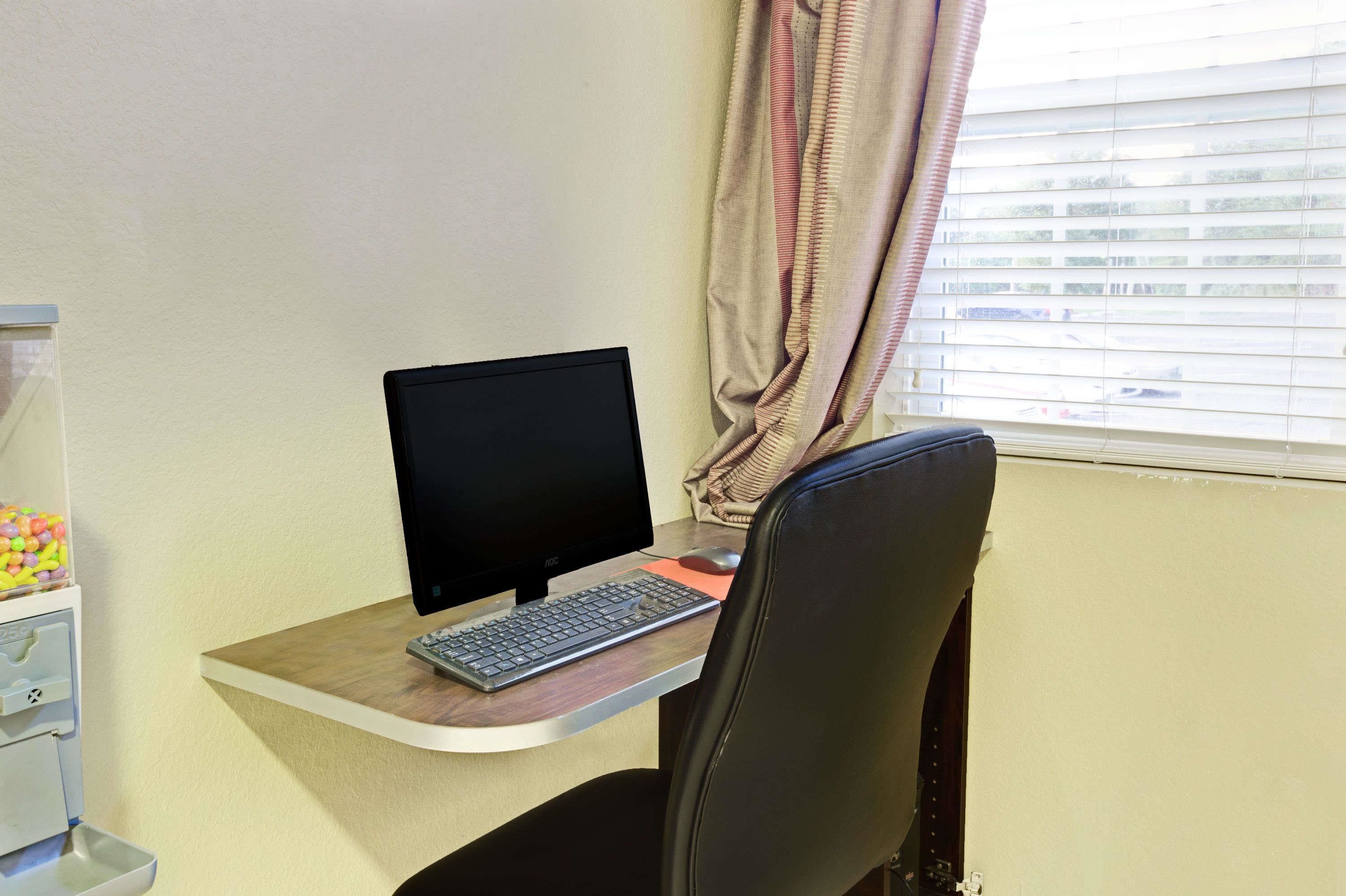A computer desk with a monitor, keyboard, and mouse. There is a chair in front of the desk. A window with blinds is visible in the background.