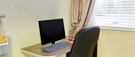 Ein Computertisch mit Bildschirm, Tastatur und Maus. Vor dem Tisch steht ein Stuhl. Im Hintergrund ist ein Fenster mit Jalousien zu sehen.