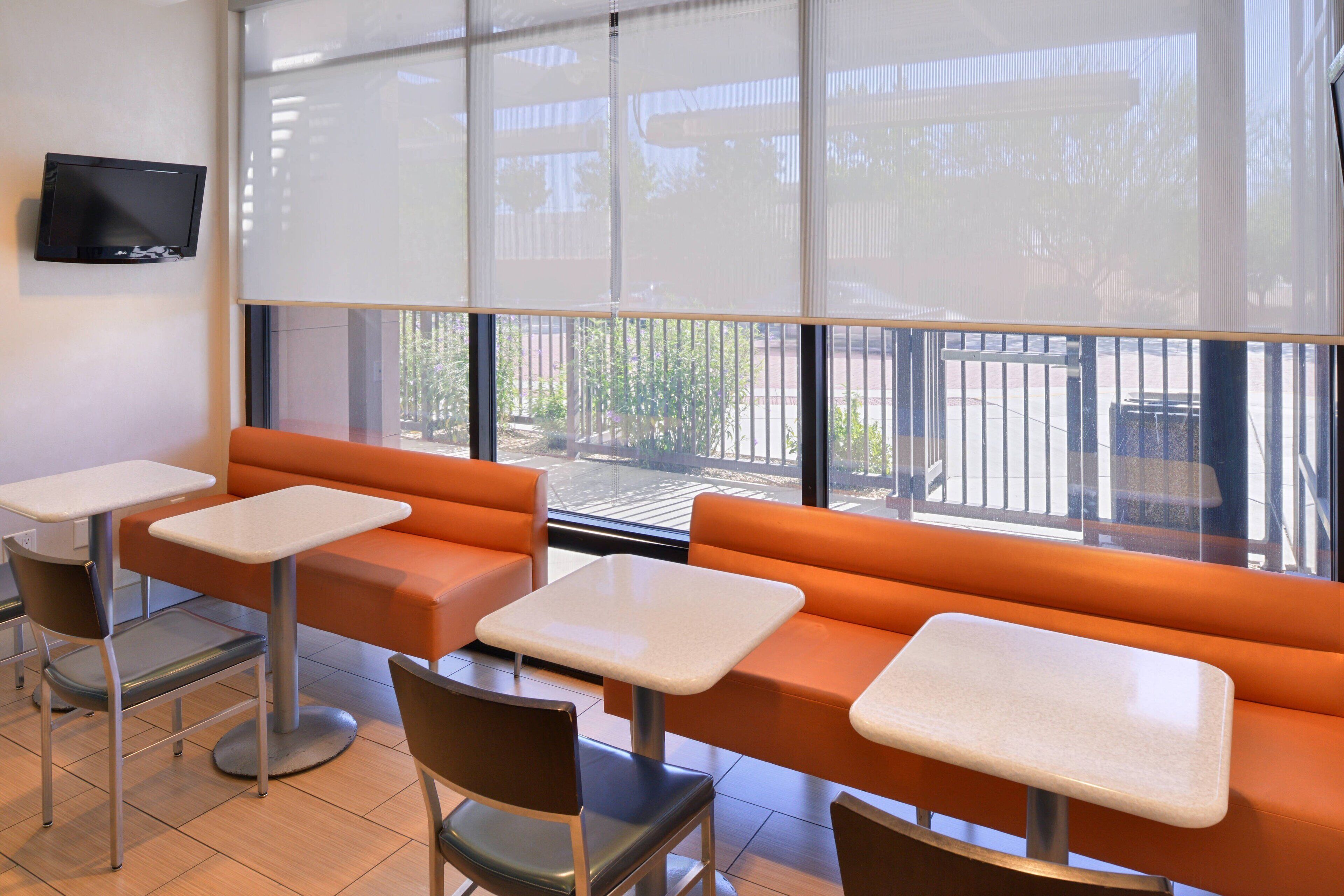 A seating area with orange booths and white tables, a television on the wall, and a view of the outside through large windows.