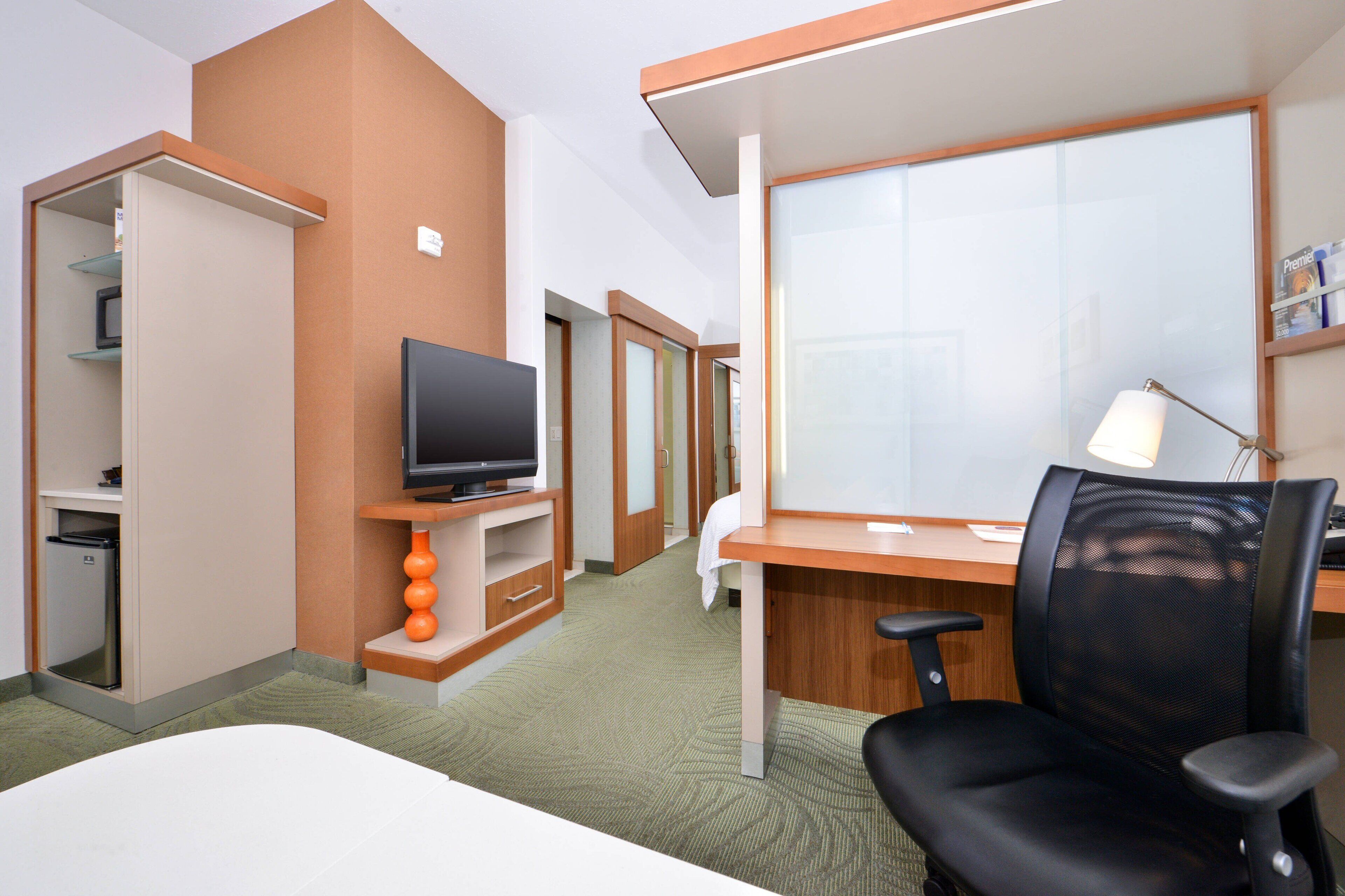 A hotel room with a bed, a desk with a chair, a television, a desk lamp, and a small kitchenette.