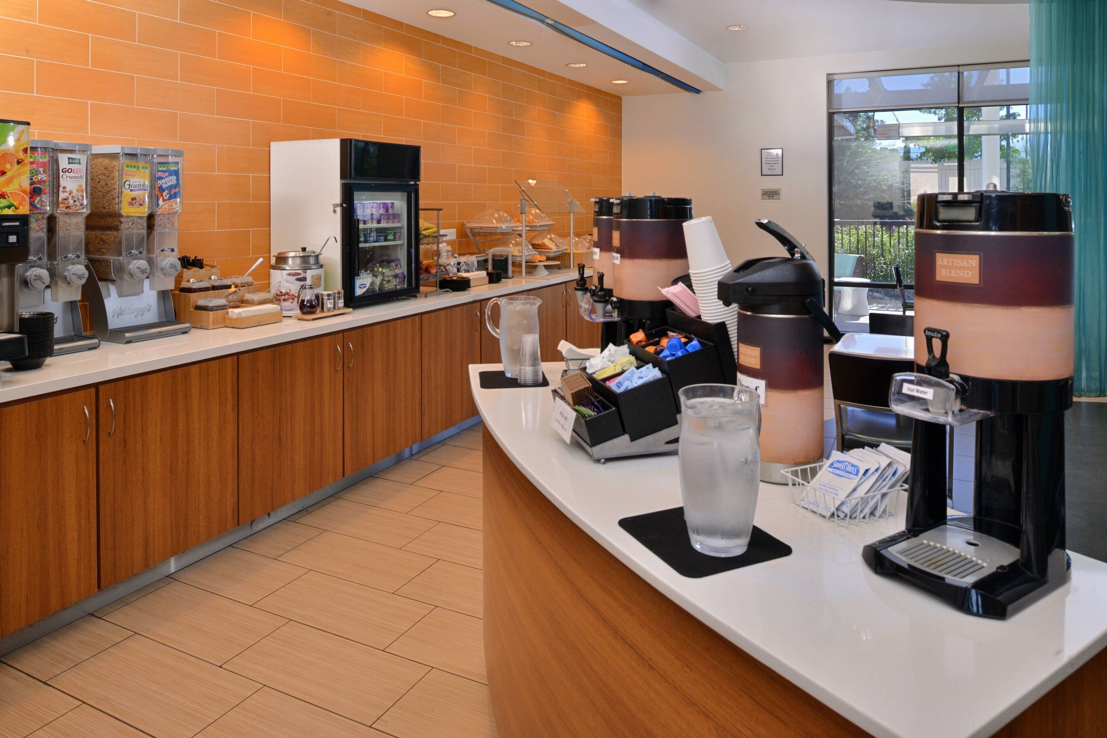 A hotel reception area with a coffee station, a refrigerator, and a window with a view of the outdoors.