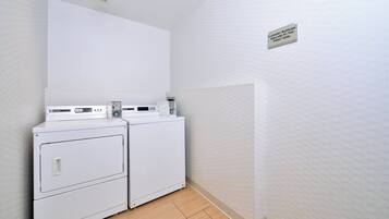 Une buanderie équipée de deux machines à laver et deux sèche-linge superposés, d’une bouche d’aération au plafond et d’un panneau indiquant l’emplacement.