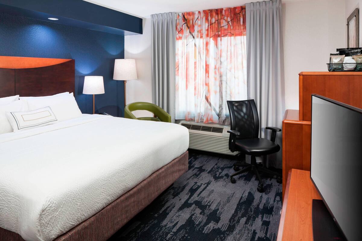 A hotel room with a bed, desk, chair, TV, and a large window with curtains.