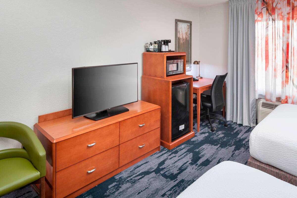 A hotel room with a flat-screen TV on a wooden dresser, a small desk with a chair, a microwave, and a refrigerator.